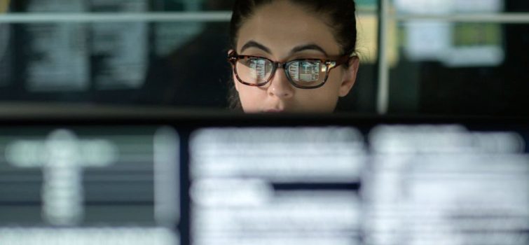 A young woman is surrounded by monitors & their reflections displaying scrolling text & data.