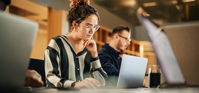 Portrait of Enthusiastic Hispanic Young Woman Working on Computer in a Modern Bright Office. Confident Human Resources Agent Smiling Happily While Collaborating Online with Colleagues.