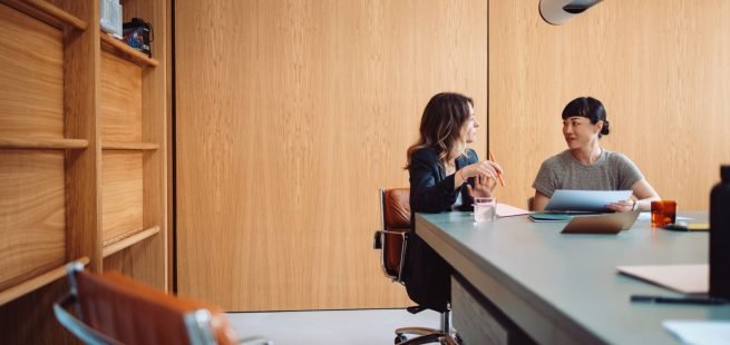 Two businesswomen engaged in a discussion at a modern office table. They are collaborating on a project with documents and laptops.